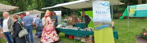 Een marktstand op een buitenbeurs met kleurrijke kleding en natuurborden op de achtergrond.