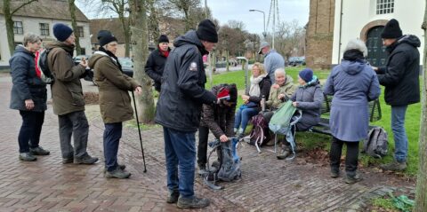 Groep ouderen met rugzakken op een plein; sommigen zitten op een bank, anderen staan.