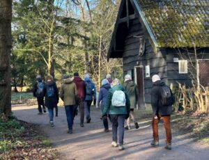 Groep wandelaars met rugzakken op bosachtig pad bij een houten huisje.