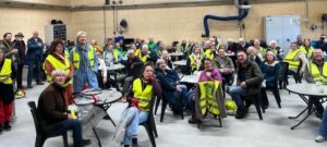 Groep mensen zit en staat in een zaal, velen dragen gele veiligheidshesjes.