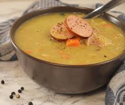 Bruine kom met erwtensoep, plakjes worst, stukjes wortel en een lepel op een marmeren ondergrond.