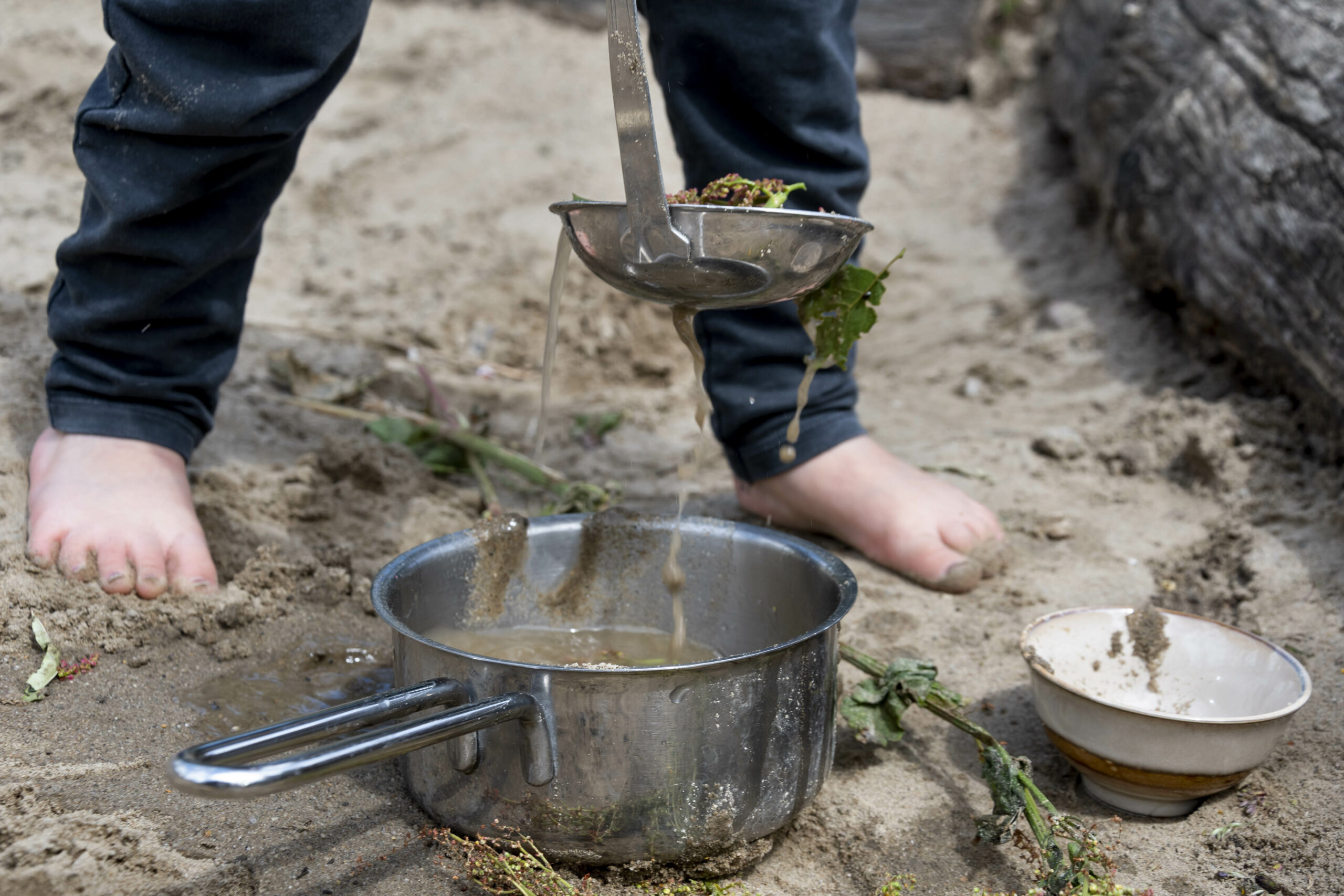 Een kind giet modder met een soeplepel in een pan buiten, met blote voeten in het zand.