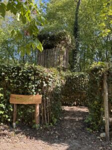 Ingang van een doolhof met houten bord omgeven door groene struiken en bomen.
