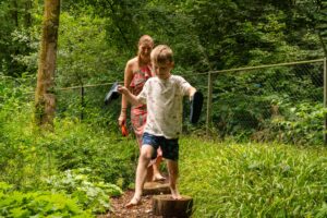 Kind en volwassene lopen op boomstammetjes in een bosrijke omgeving, beiden dragen schoenen in hun handen.