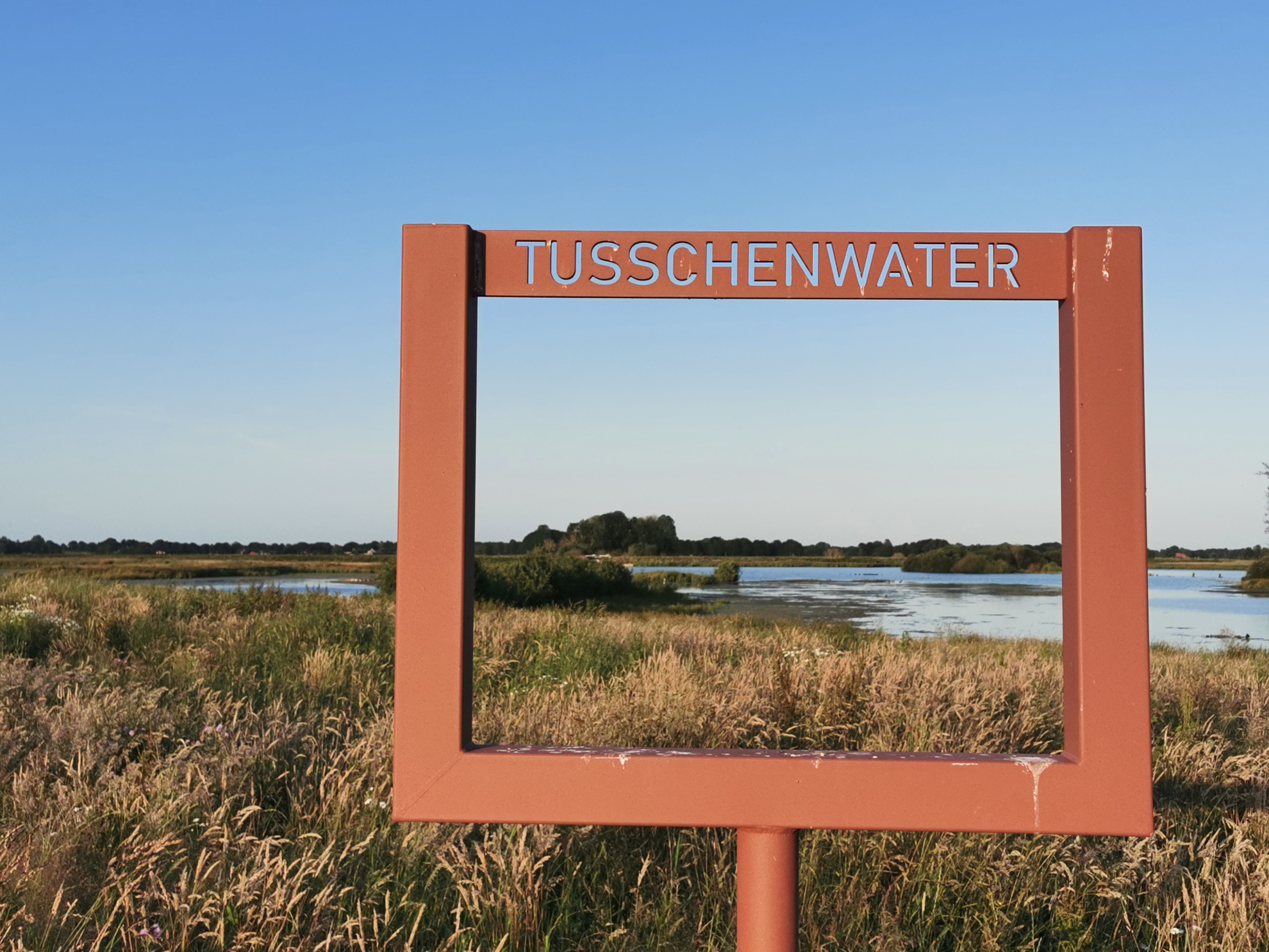 Rood frame met "TUSSCHENWATER" omkadert een graslandschap met water en blauwe lucht.
