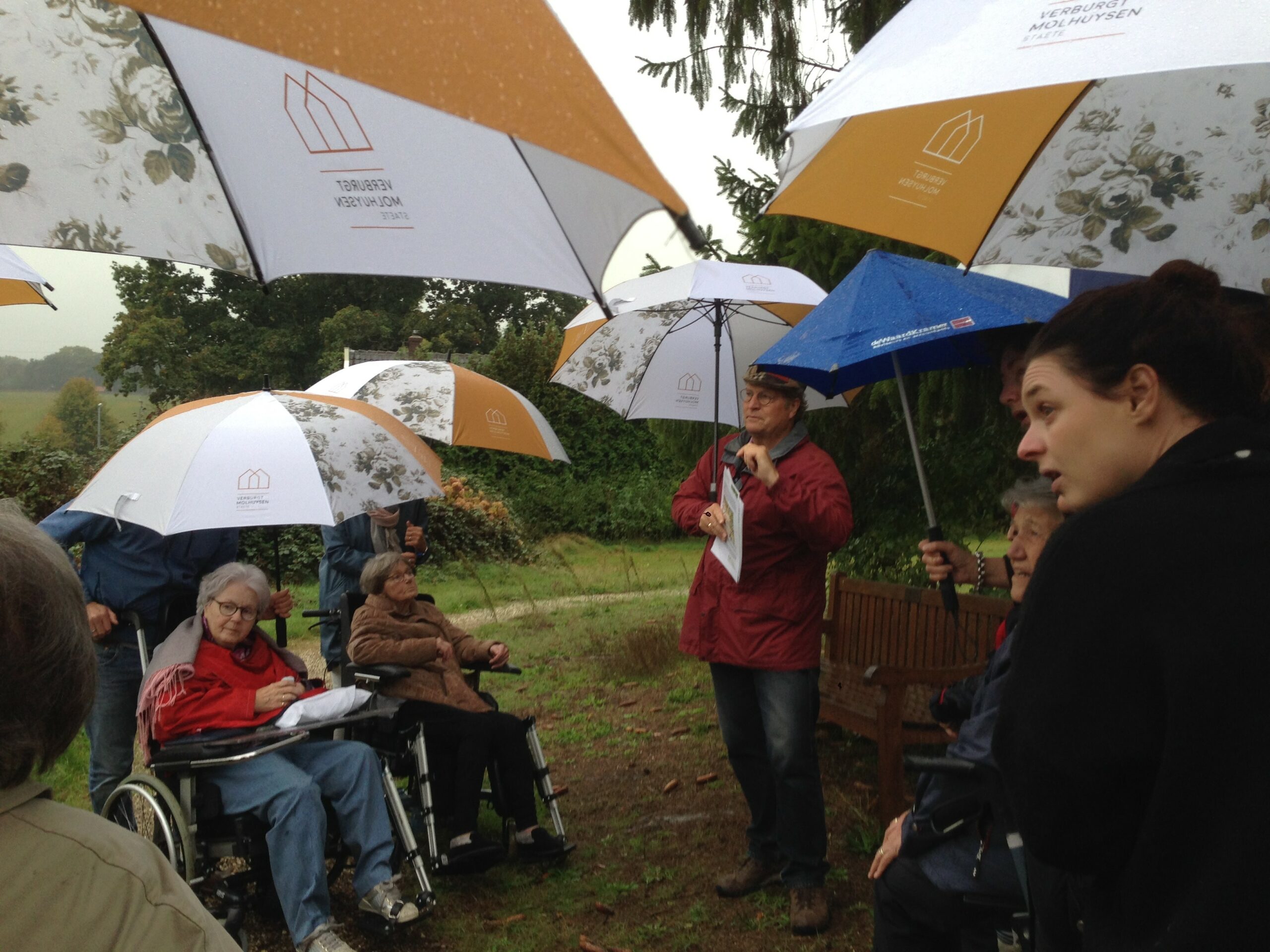 Groep mensen met paraplu's, enkele in rolstoelen, buiten in de regen.
