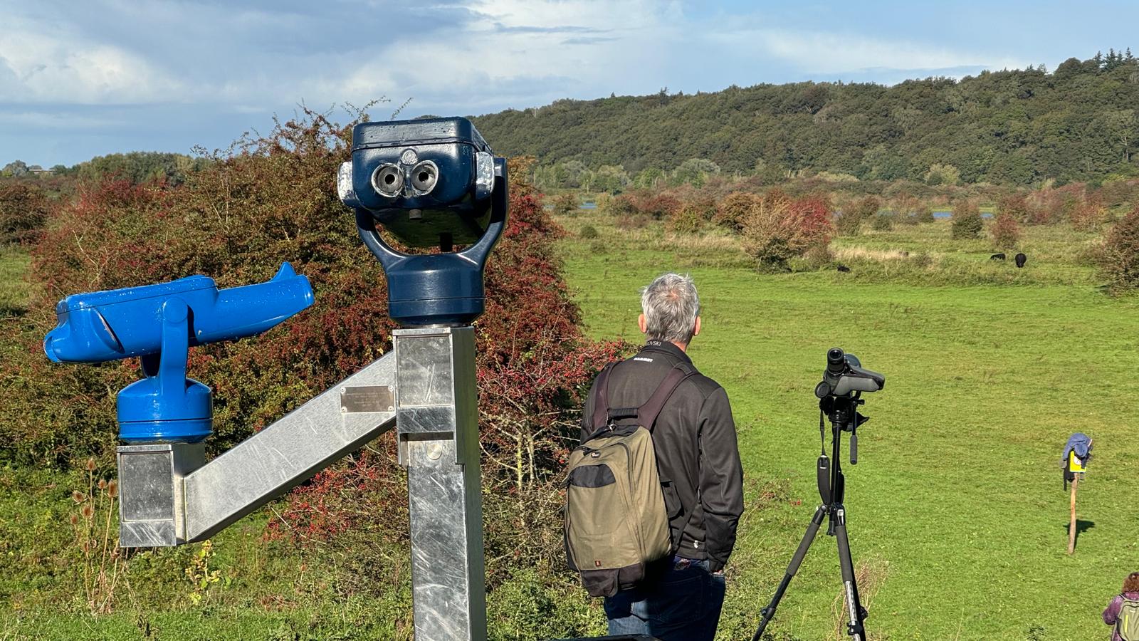 Man met rugzak kijkt naar groene weide met verrekijker en blauwe telescoop.