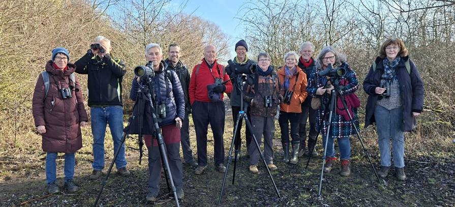 Groep vogelaars met camera's en verrekijkers poseren buiten bij een bosrijke achtergrond.