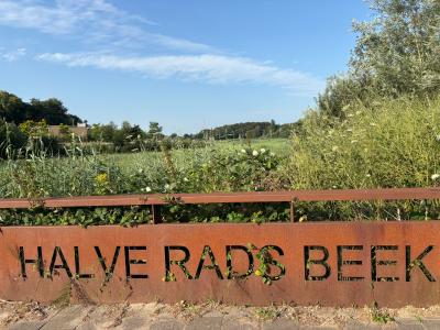 Roestige metalen sign met tekst "HALVE RADS BEEK" voor een weelderig groen landschap.