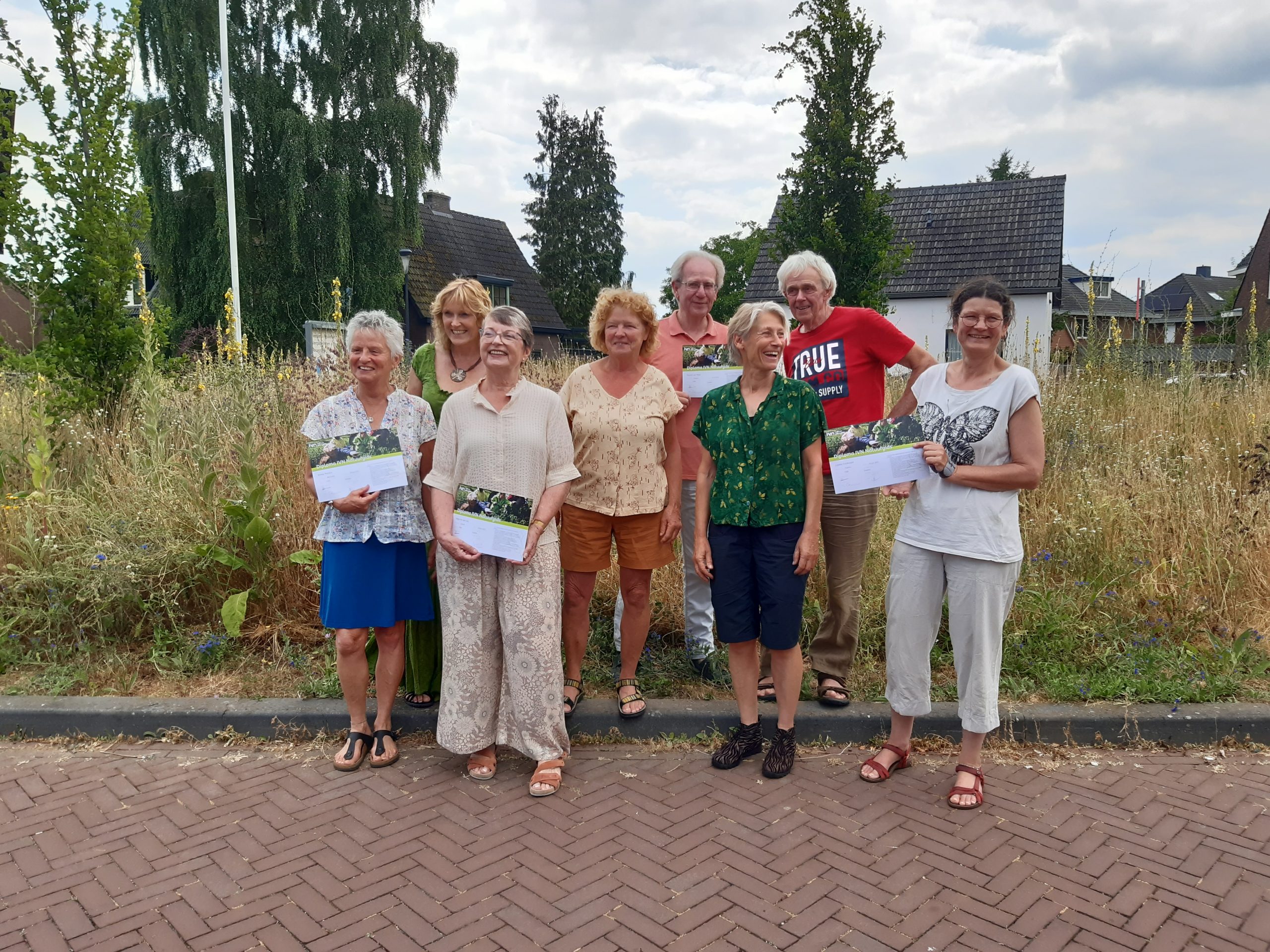 Een groep mensen poseert met certificaten voor een groene achtergrond in een tuinwijk.