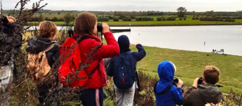 Kinderen met verrekijkers kijken uit over een rivierlandschap met een auto aan de oever.