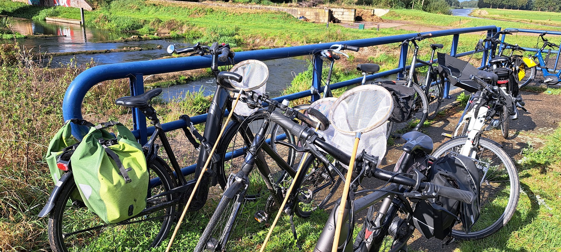 Fietsen met zadeltassen en schepnetten staan tegen een blauwe reling bij een rivier.