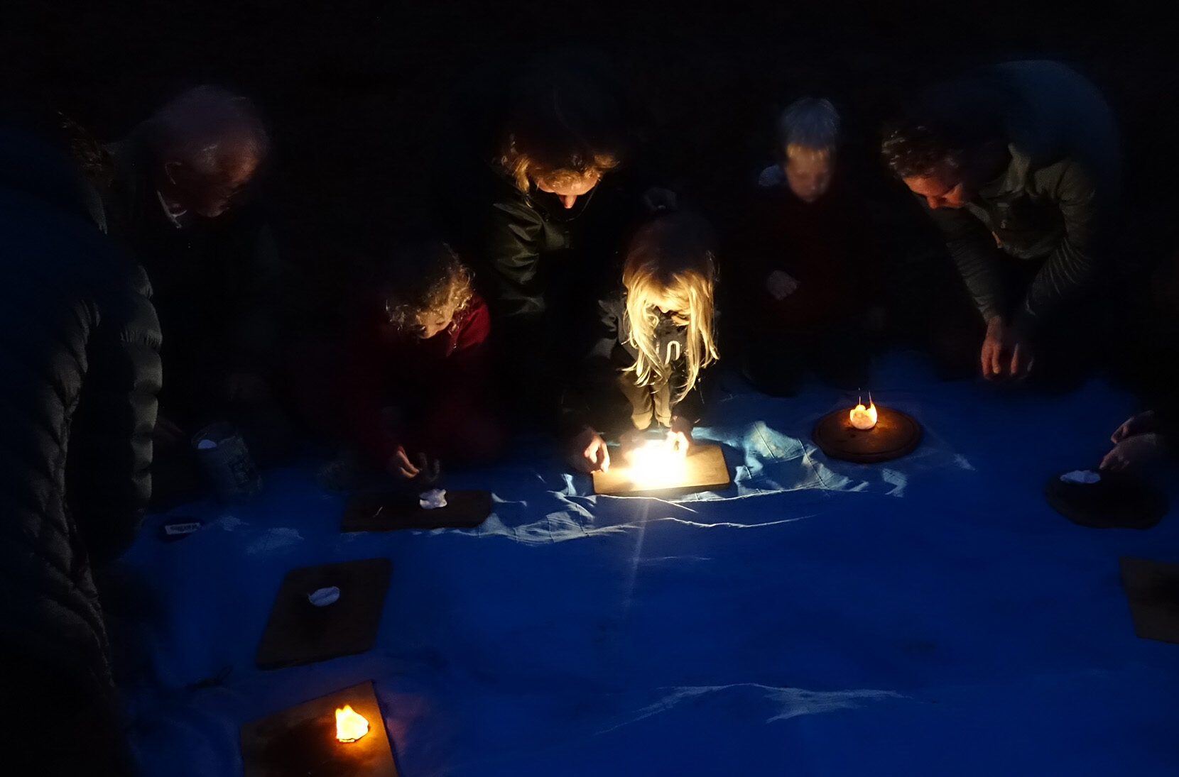 Mensen rond een kaarslicht zittend op een blauwe doek in het donker.