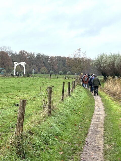 Mensen wandelen op een smal pad langs een groen weiland met houten hek en een witte poort in de verte.