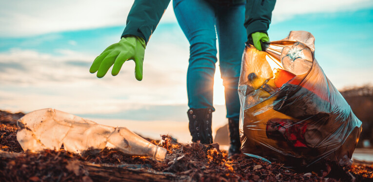 Persoon verzamelt afval op strand met vuilniszak en handschoenen, bij ondergaande zon.