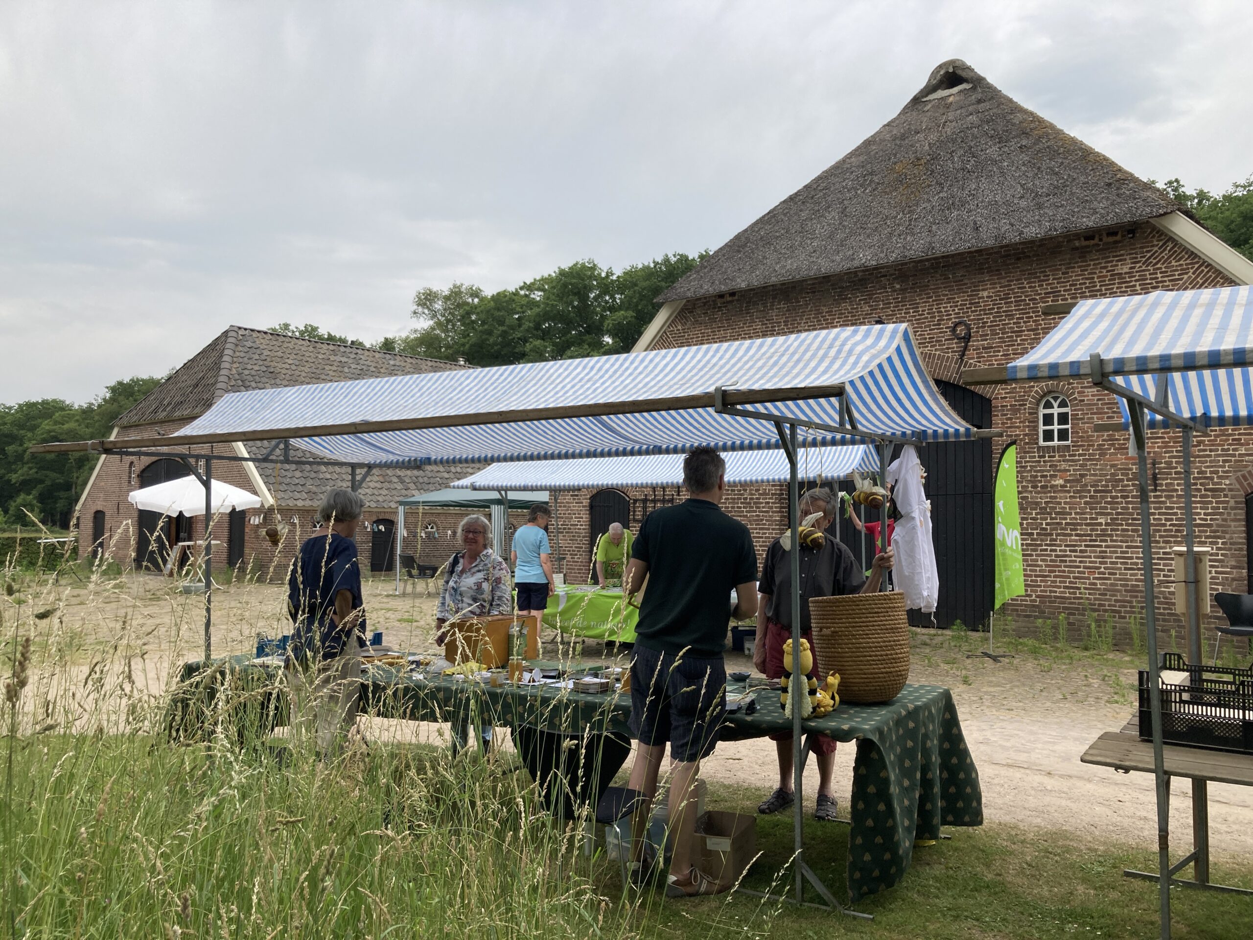 Marktkramen met blauw-witte luifels voor een oude schuur, omringd door gras, mensen converseren.