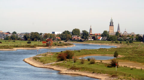 Rivierlandschap met een dorp, windmolen en kerktorens op de achtergrond.