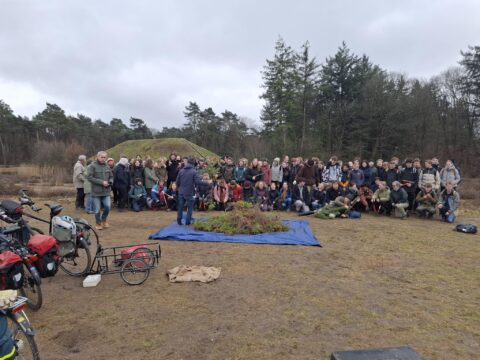 Grote groep mensen poseert buiten bij een groen bedekte heuvel, omringd door bos.