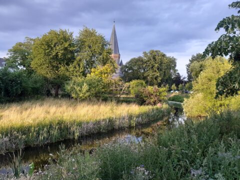 Landschap met kerkspits, bomen en een kronkelende beek onder een bewolkte hemel.