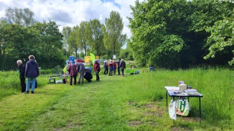 Een groep mensen verzamelt zich op een grasveld bij een tafel met boeken en bomen op de achtergrond.