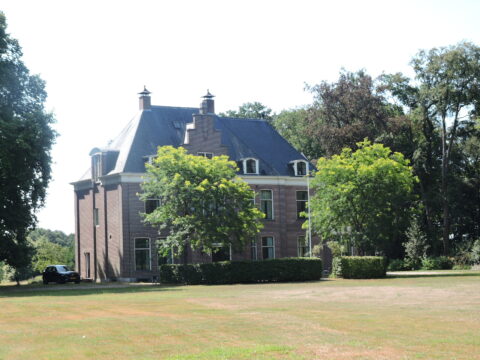 Oud herenhuis met een omringend grasveld en bomen op een zonnige dag.