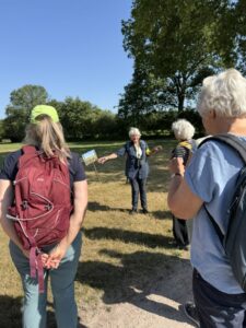 Een groep mensen luistert naar een gids in een zonnige, groene omgeving met bomen.
