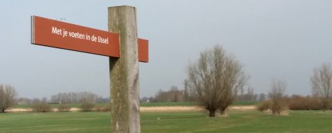 Brug met bord "Met je voeten in de IJssel" in een open landschap met bomen en gras.