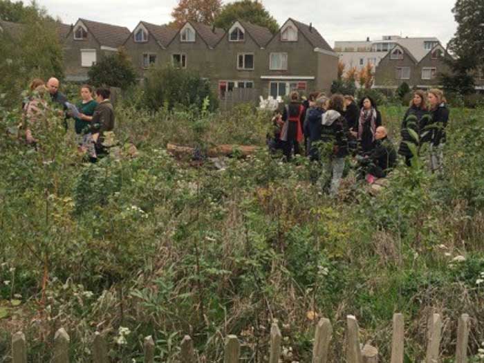 Groep mensen staat en praat in een groene, overwoekerde tuin met huizen op de achtergrond.