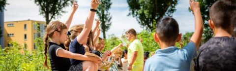 Kinderen buiten steken hun handen op, omringd door groen en gebouwen.