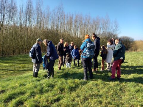 Groep mensen op wandeling in groen veld met kale bomen op achtergrond, in zonnig weer.
