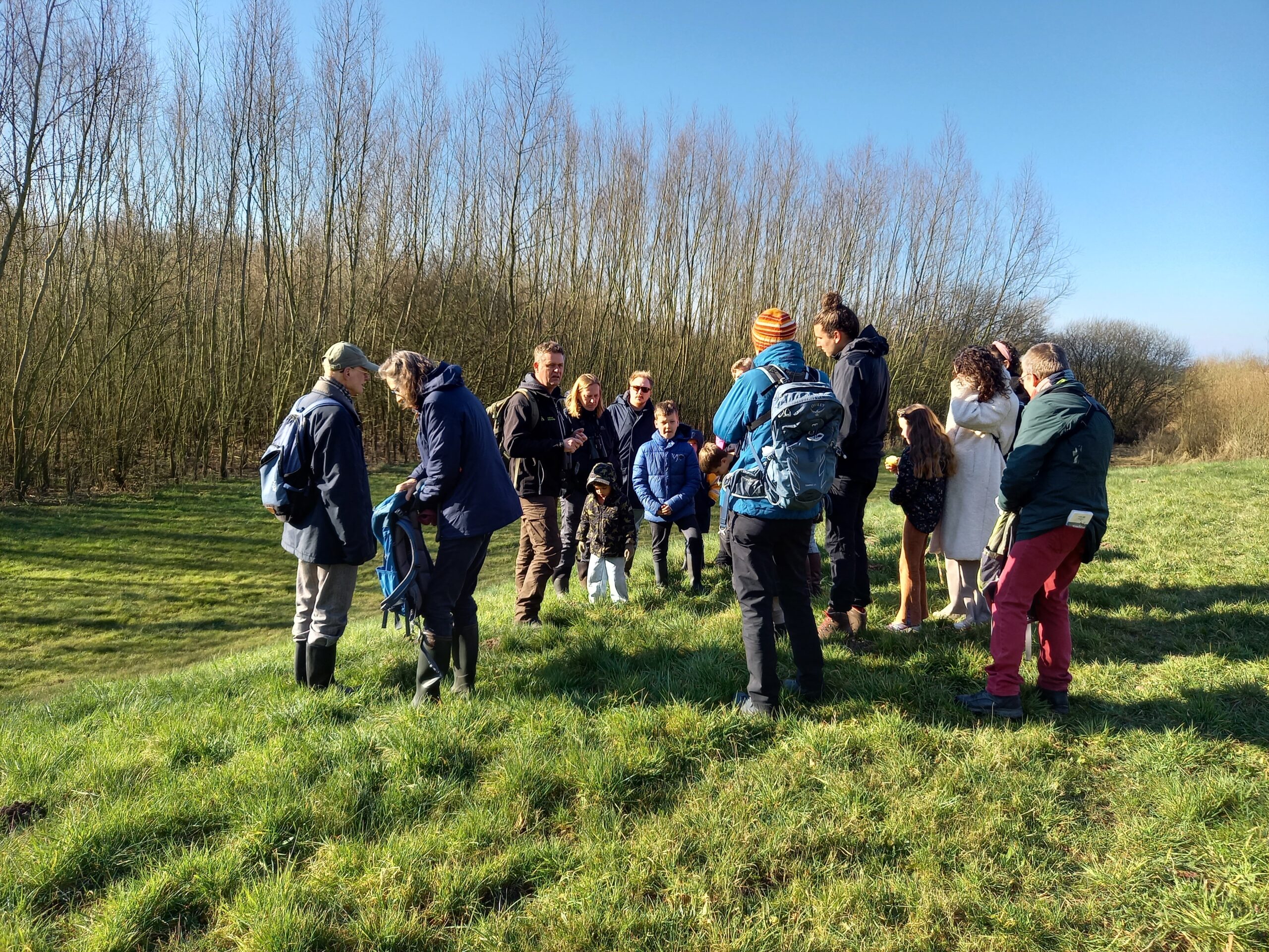 Groep mensen op wandeling in groen veld met kale bomen op achtergrond, in zonnig weer.
