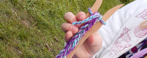 Hand houdt houten vork vast met gevlochten paars en blauw garen, gras op de achtergrond.