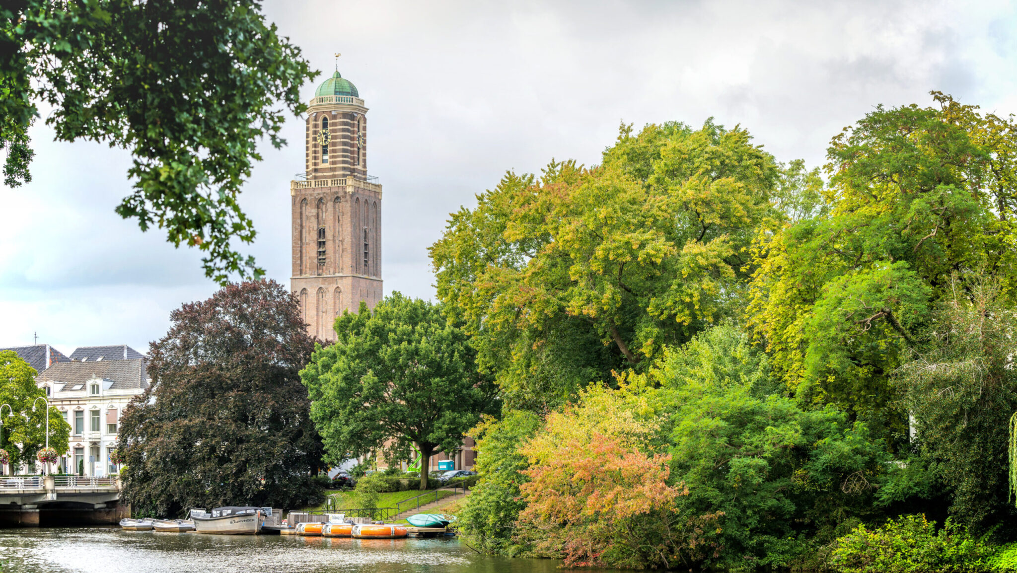 Toren met groene koepel naast bomen en rivier; boten aangemeerd bij oever.