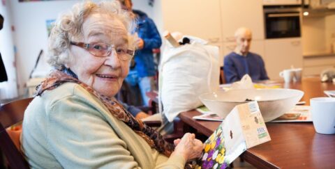 Een lachende oudere vrouw houdt een zakje bloemzaden vast aan een tafel in een huiskamer.