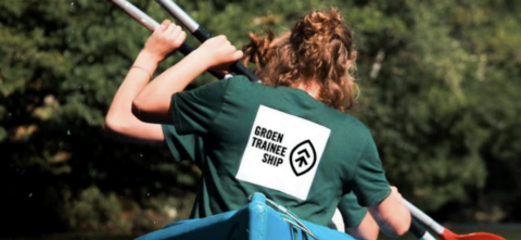 Twee personen in een kano dragen shirts met "Groen Traineeship" op een bosrijke achtergrond.