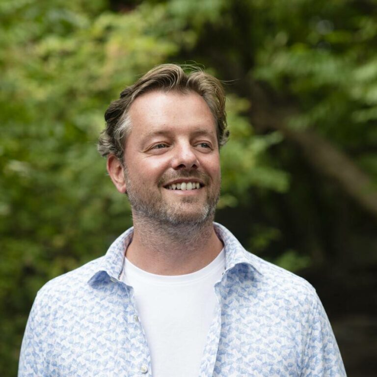 Man glimlacht buiten met bomen op de achtergrond, draagt een wit shirt en lichtblauw overhemd.