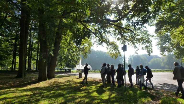 Groep mensen in een zonnig bos observeert een fontein op de achtergrond.