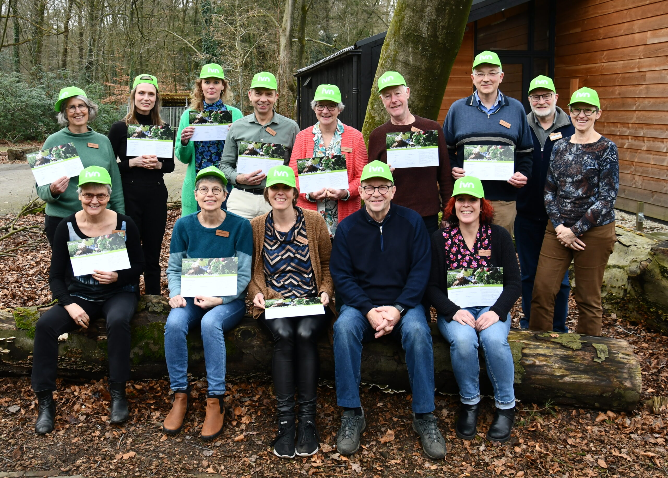 Groep mensen met groene petten, houdt certificaten vast bij een boom in een bosrijke omgeving.