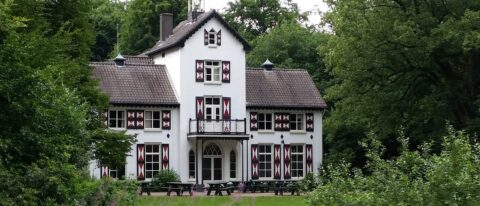 Een wit landhuis met rood-witte luiken omgeven door groen en bomen.