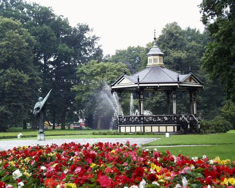 Bloemenperk met standbeeld op de voorgrond en prieel met fontein in groene parkachtergrond.
