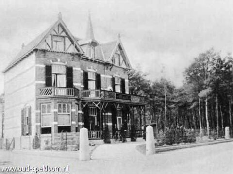 Oud, statig herenhuis met houten veranda, omringd door bomen en hekwerk.