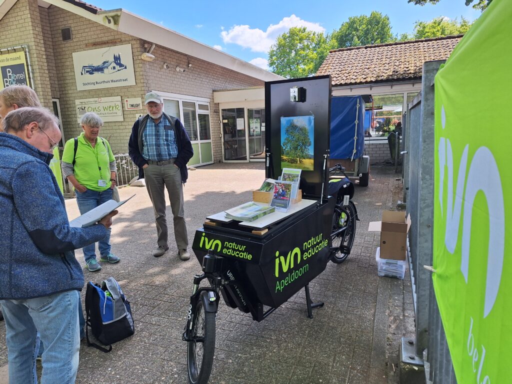 Groep mensen bij IVN-promotiekraam met natuurposters buiten gemeenschapshuis.