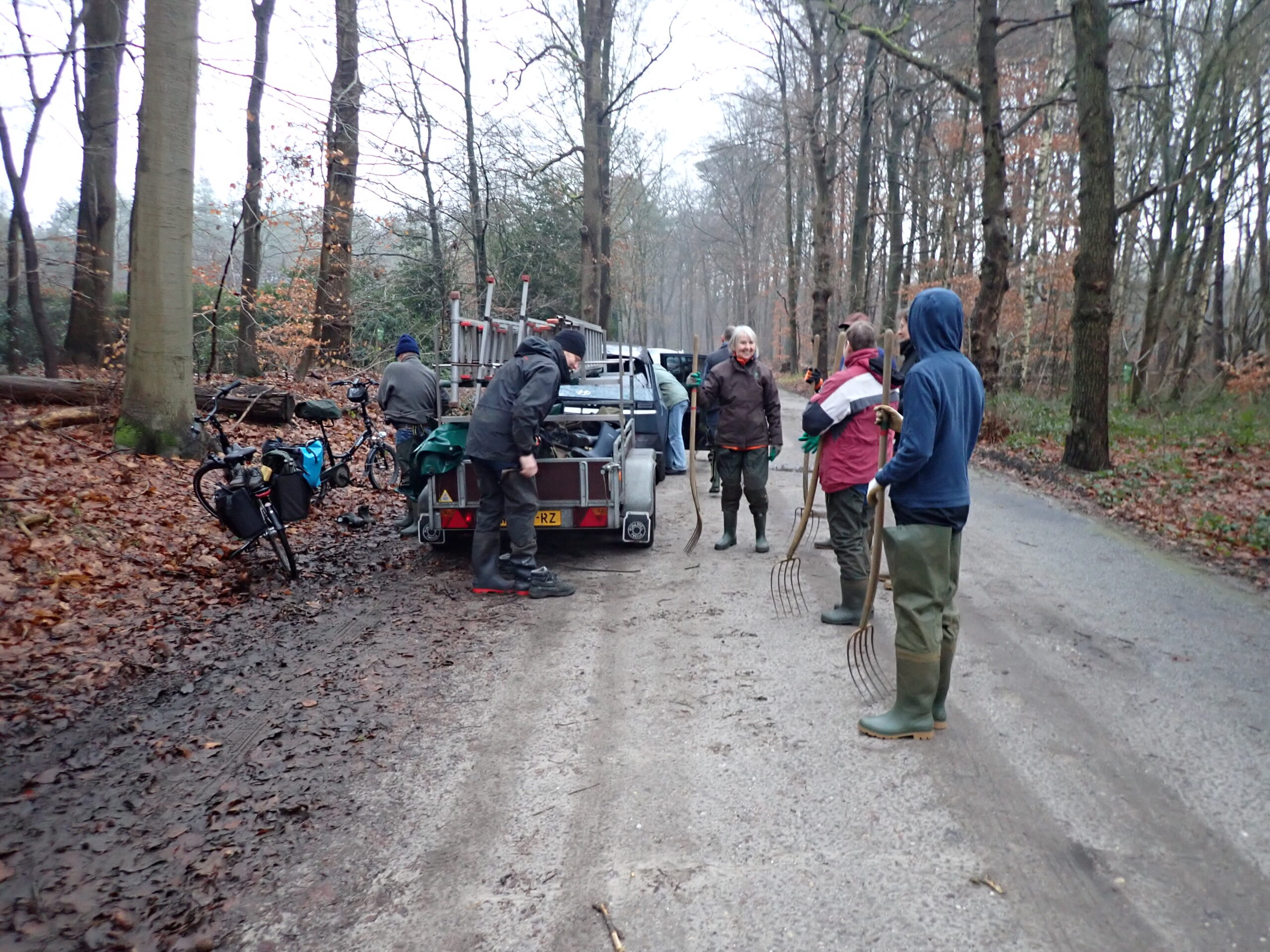 Mensen verzamelen met harken rond een auto en aanhanger in een bosrijke omgeving.