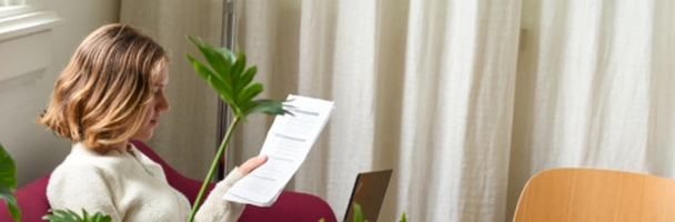 Vrouw leest een document op een stoel, omringd door planten in een kamer met gordijnen.