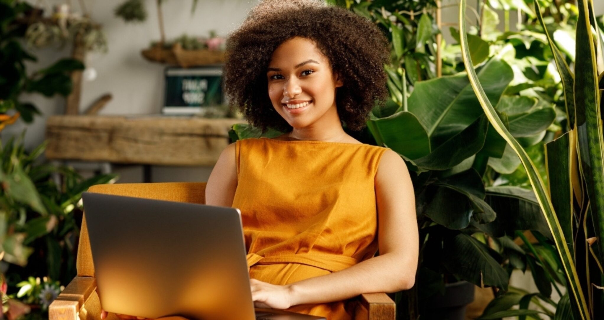 Vrouw in een oranje jurk werkt op laptop, omringd door groene planten.