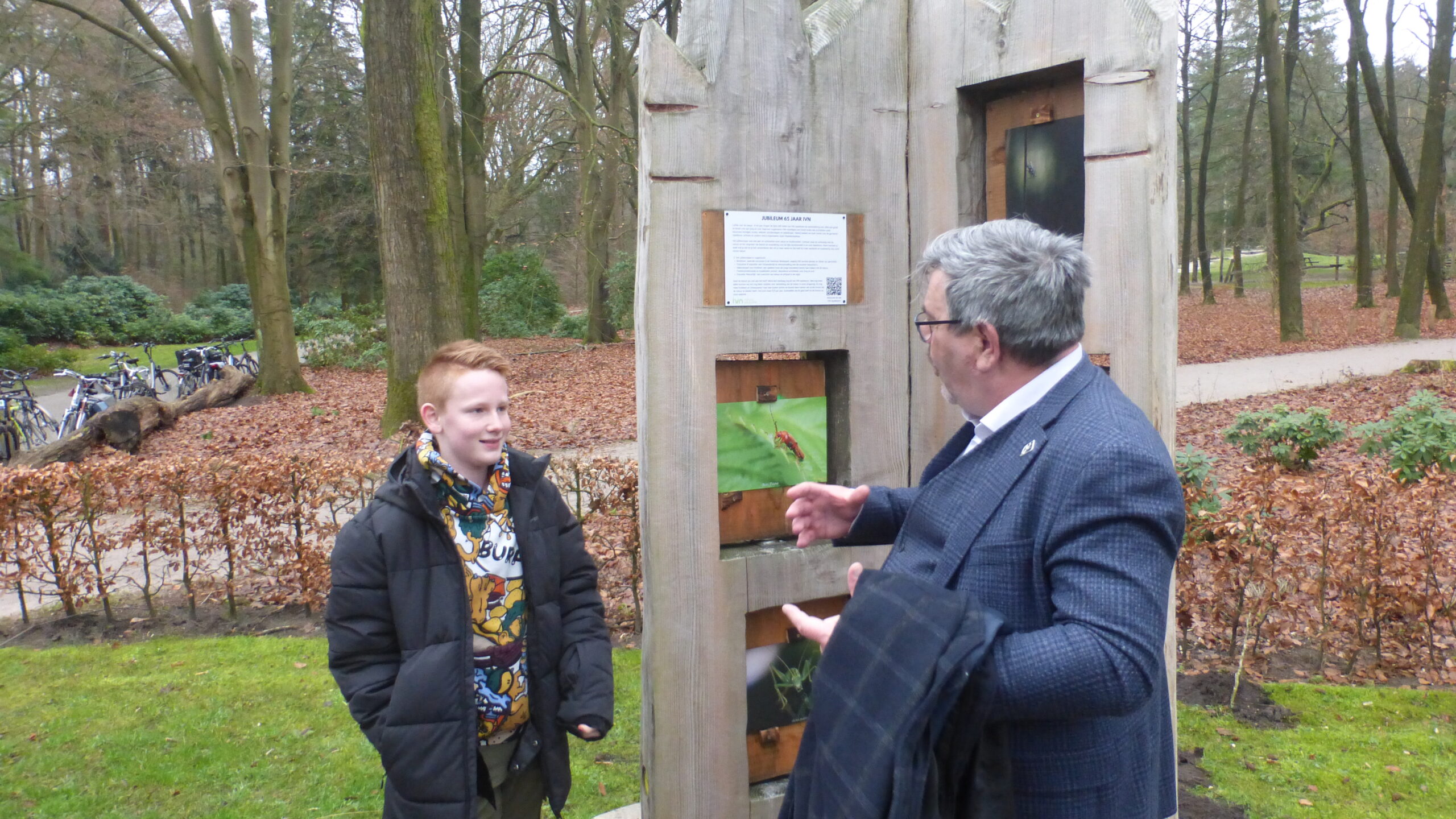 Twee mensen praten buiten bij een informatiebord in een bosrijke omgeving.