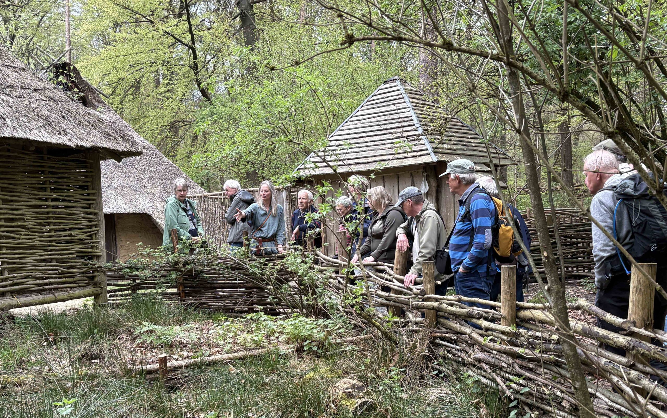 Groep mensen bij historische strodaken in bosrijke omgeving met houten omheining.