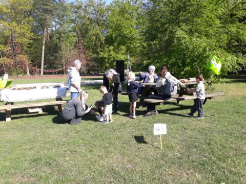 Groep volwassenen en kinderen bij picknicktafel in park, met op de achtergrond speeltuin en bomen.