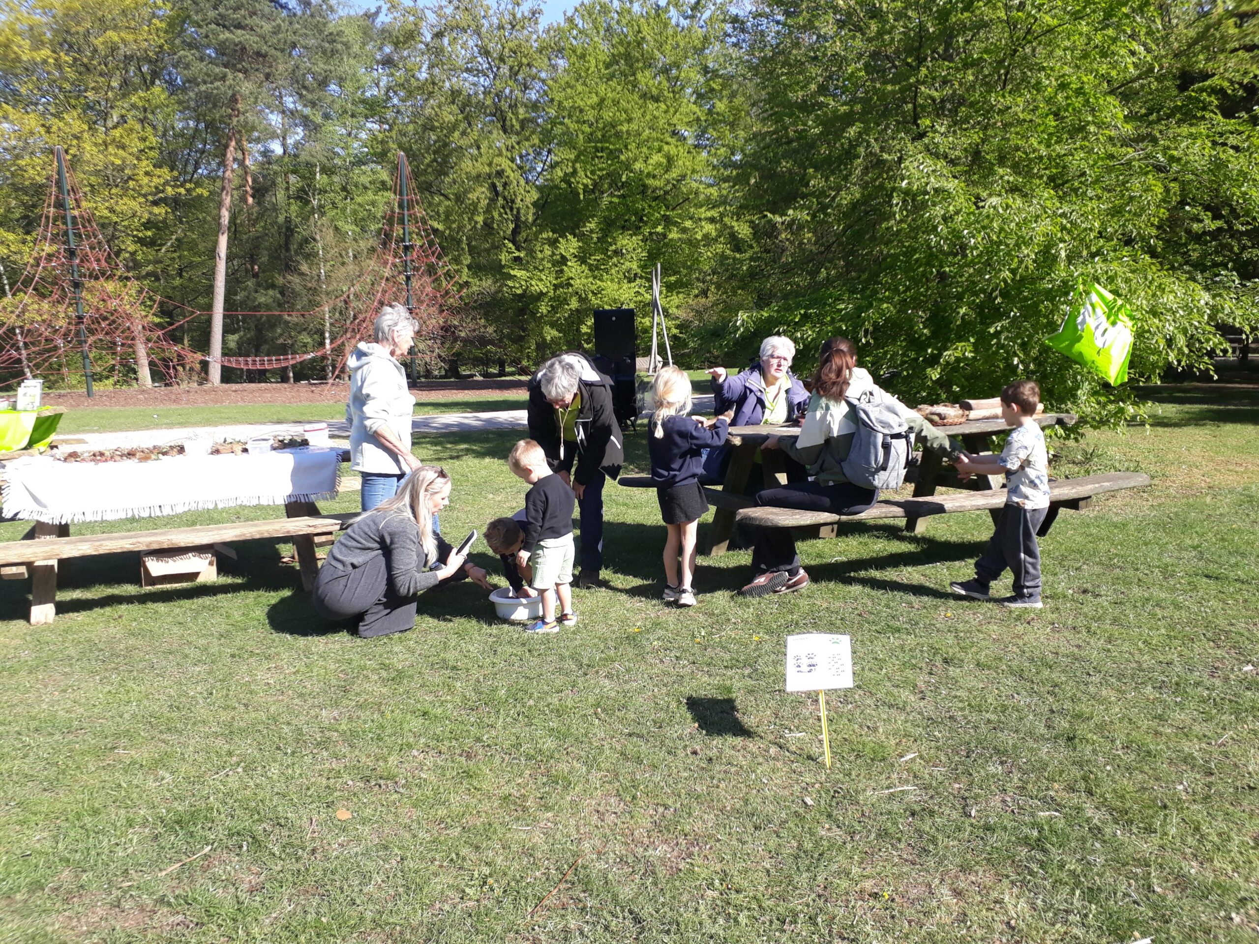 Groep volwassenen en kinderen bij picknicktafel in park, met op de achtergrond speeltuin en bomen.