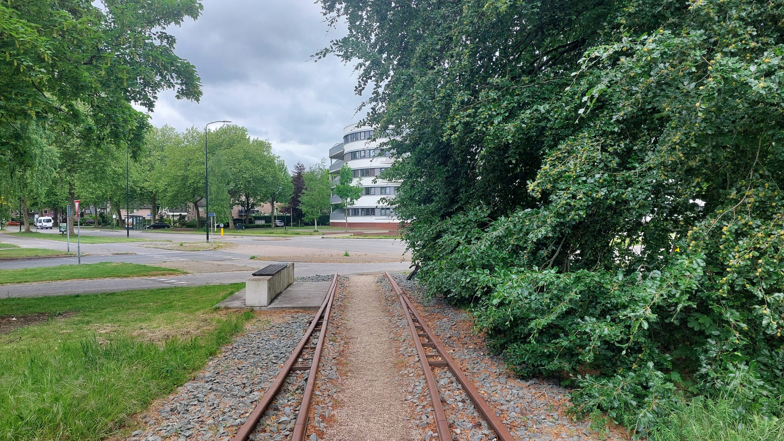 Verlaten smalspoor spoorweg met stenen en gras, omringd door bomen en een modern gebouw op de achtergrond.